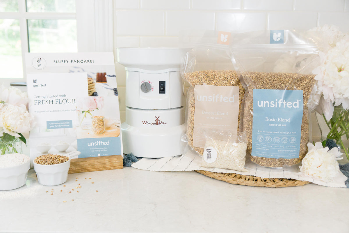 WonderMill Electric Grain Mill shown with the Unsifted Starter Kit: Unsifted Basic Blend and Dessert Blend grain bags, Recipe Cards, the Unsifted Prime Pack and the Getting Started Guide